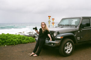 Solo female traveller holding her camera leaning on a jeep while road tripping through Hawaii. Background is the ocean.
